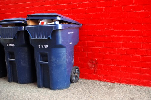 Two large dark blue plastic rubbish bins with closed lids positioned on a concrete pavement next to a bright red brick wall in an outdoor setting. The bins have textured surfaces with vertical ridges and visible wheel mechanisms on one side, with one bin slightly overlapping the other. The lid of the bin on the left is partially open, revealing some waste materials inside. The background includes a clean, well-maintained red brick wall, indicating a commercial or residential exterior environment. The lighting is natural, casting soft shadows on the pavement and highlighting the texture of the bins and the brickwork, reflecting typical rubbish removal collection points used by companies such as Commercial Waste Removal Harrow in the Harrow area. The scene captures a moment in a waste management process aimed at local rubbish clearance within the region, emphasizing the practical storage of waste awaiting collection.