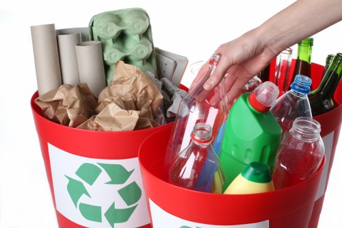 A close-up view of two red recycling bins with white labels displaying the recycling symbol, placed outdoors on a paved surface, filled with various waste items. The left bin contains several cardboard tubes and crumpled brown paper, while the right bin is filled with multiple plastic bottles, including clear, green, and blue ones, some with caps on, and a few other plastic containers. A person's hand is reaching into the right bin, level with the top of the bottles, suggesting active disposal. The environment appears to be a typical urban or suburban setting, possibly near a commercial or residential building, indicative of waste collection or rubbish removal activities in the Harrow area. The overall scene emphasizes waste segregation and proper disposal practices aligned with rubbish removal services provided by Commercial Waste Removal Harrow.