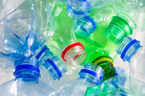 A close-up image of numerous used plastic bottles with multicoloured caps, including blue, green, and red, piled together on a reflective surface. The bottles are transparent, showing their empty interior, with some slightly crushed or deformed. The caps are pressed tightly onto the bottles, and the plastic's texture appears smooth and glossy. The background is indistinct, but the overall scene suggests a focus on plastic waste, relevant to rubbish removal services such as those offered in Harrow by Commercial Waste Removal Harrow, highlighting the importance of proper disposal of recyclable plastic materials.