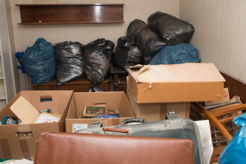 The image shows a cluttered indoor storage area or room with several large black and blue rubbish bags stacked against a wooden wall, indicating waste accumulation from rubbish removal activities. In the foreground, there are open cardboard boxes filled with various miscellaneous items, possibly paper, packaging, or small discarded objects. A portable vacuum cleaner or similar device is partially visible, placed next to the boxes. To the right, there is a wooden slatted structure or rack, and the floor appears to be covered with a brown material, possibly carpet or tile. The overall scene suggests an environment where waste or waste-related items are temporarily stored or prepared for removal, consistent with commercial waste removal services such as those offered by Commercial Waste Removal Harrow, serving Harrow and nearby areas. Lighting is even, with some shadows cast by the objects, emphasizing a practical, work-in-progress setting related to rubbish collection or disposal activities.