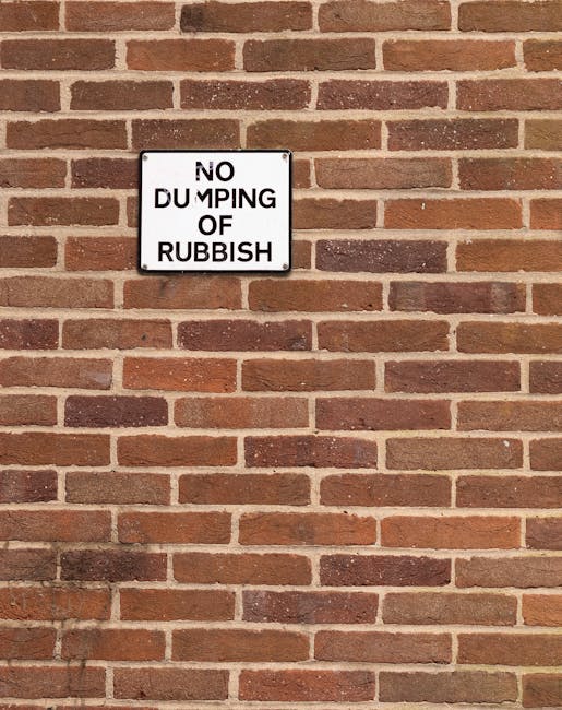 A rectangular metal sign mounted on a brick wall with a white background and black text that reads 'NO DUMPING OF RUBBISH'. The wall is constructed of reddish-brown bricks with a consistent horizontal