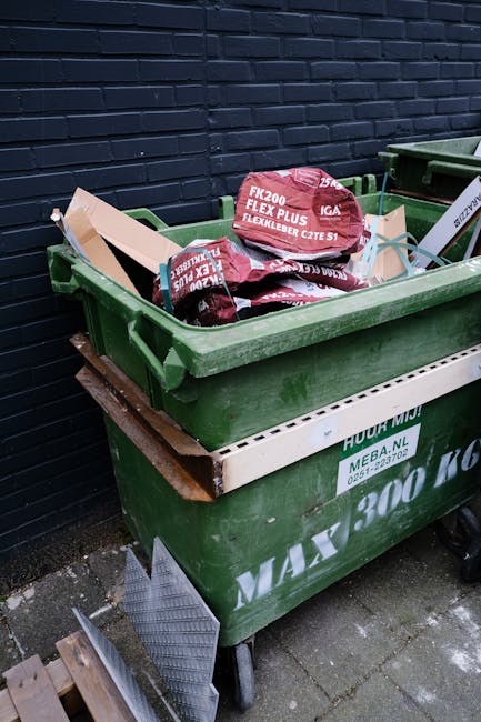 A close-up view of a waste collection site showcasing a mixed assortment of rubbish including black plastic bags, a yellow plastic container, an old, dirty car tire leaning on a pile of waste, and var