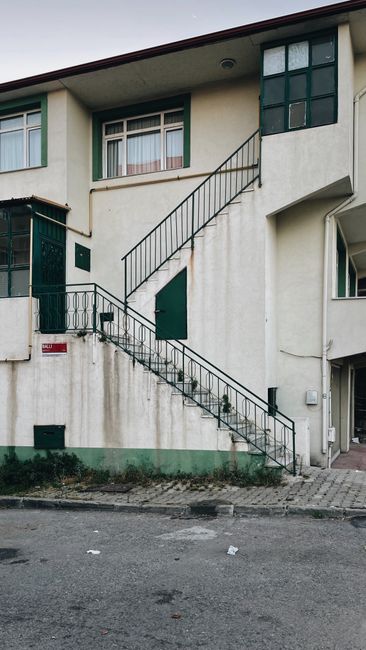 A concrete outdoor staircase attached to the side of a building, with visible signs of weathering and graffiti marks on the lower section. The staircase has a black metal railing along the sides, and 